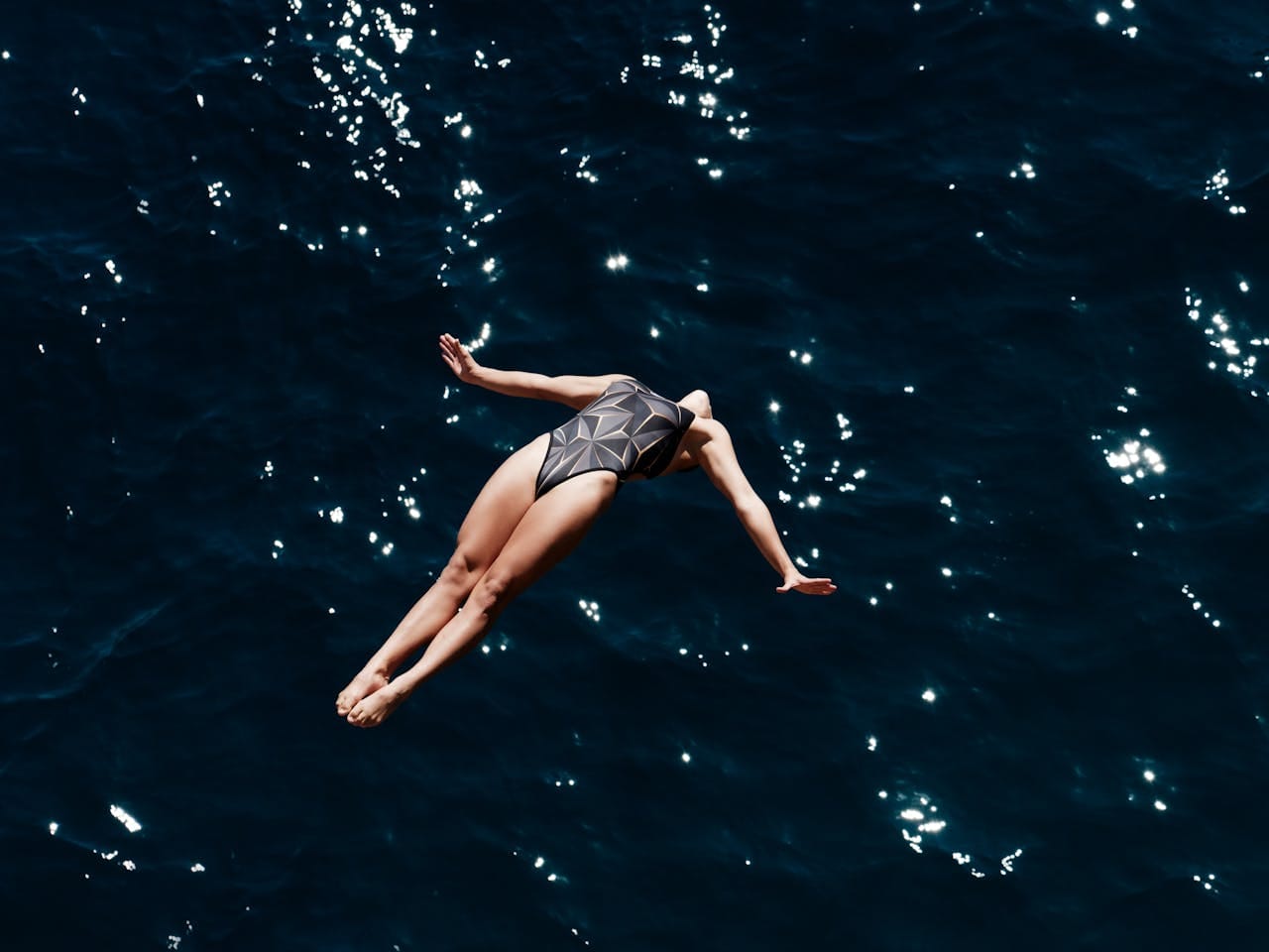 A person diving into dark water with glittering light reflections, wearing a geometric patterned swimsuit, arms outstretched in a graceful pose.